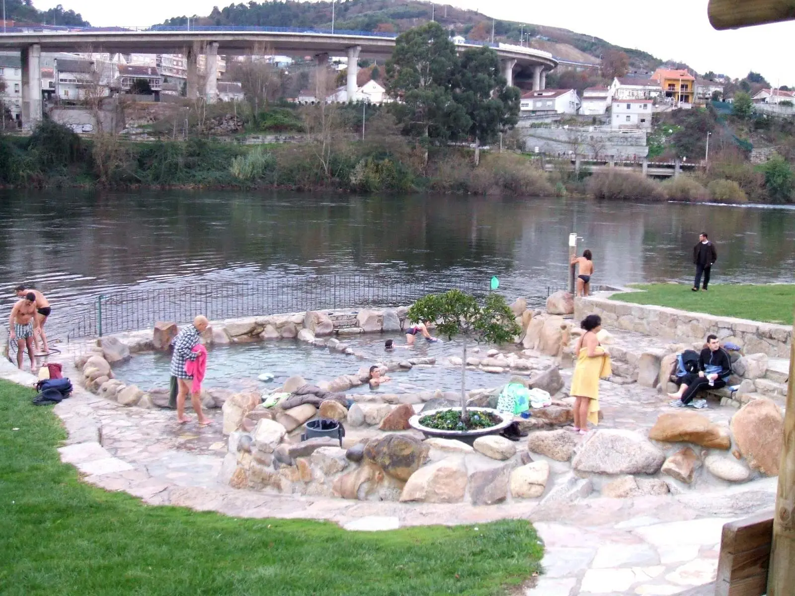 Un bassin rond aux bords de galets cimentés, au ras du Miño, où quelques personnes se baignent ou se sèchent sur les dalles, avec une pelouse au premier plan et un viaduc sur l’autre rive.