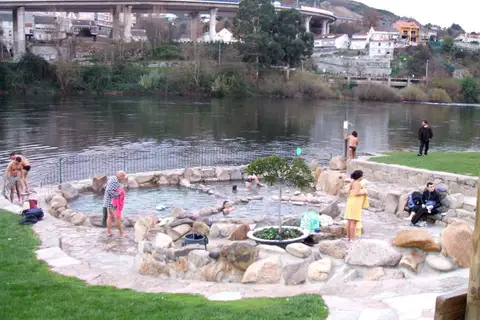 Un bassin rond aux bords de galets cimentés, au ras du Miño, où quelques personnes se baignent ou se sèchent sur les dalles, avec une pelouse au premier plan et un viaduc sur l’autre rive.