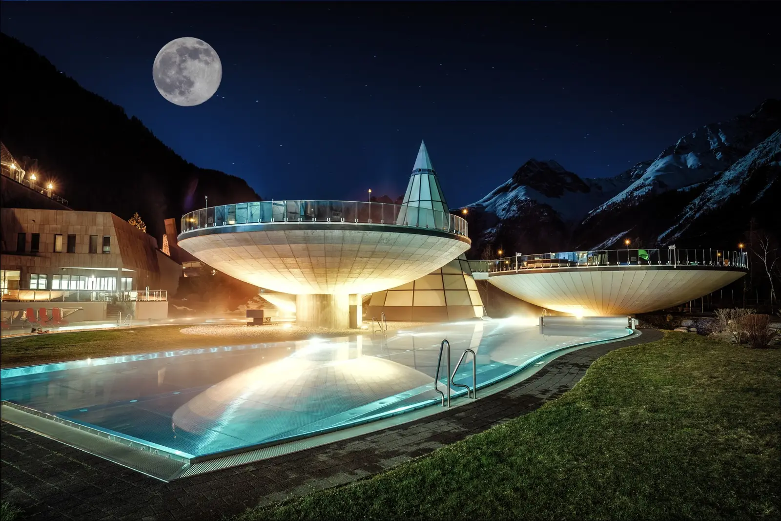 Bassins extérieurs éclairés de l’intérieur dans la nuit, le bâtiment conique de verre allumé derrière eux, sous une pleine lune et des montagnes enneigées.