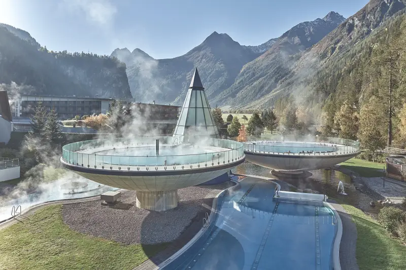 Deux bassins circulaires surélevés, fumants, posés sur des socles en béton devant un bâtiment de verre en forme de cône, au fond d’une vallée alpine boisée aux sommets encore dégagés.
