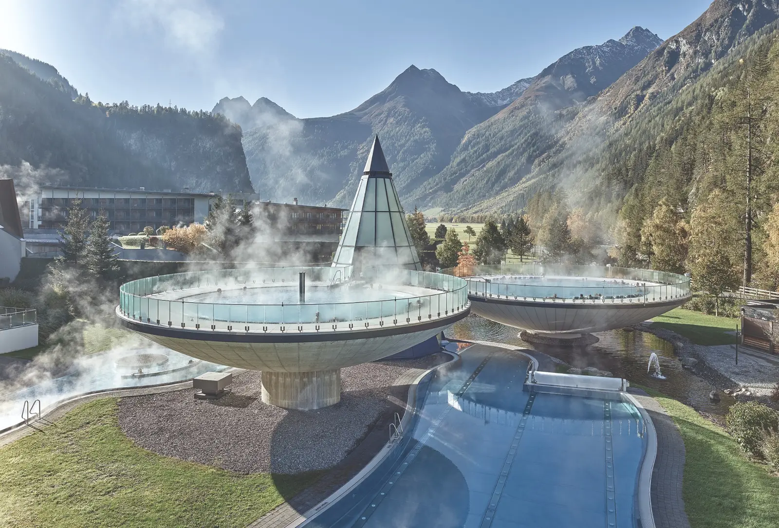 Deux bassins circulaires surélevés, fumants, posés sur des socles en béton devant un bâtiment de verre en forme de cône, au fond d’une vallée alpine boisée aux sommets encore dégagés.