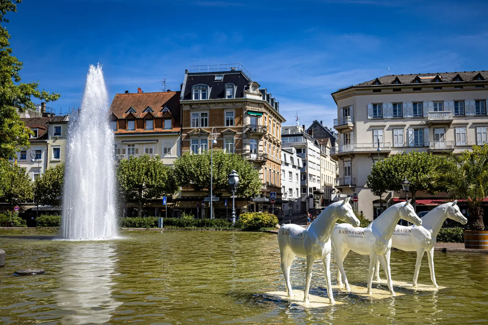 Trois chevaux sculptés blancs, portant l’inscription « BADEN-BADEN », se tiennent dans un bassin du centre-ville de Baden-Baden devant un jet d’eau et des immeubles anciens, en plein été.