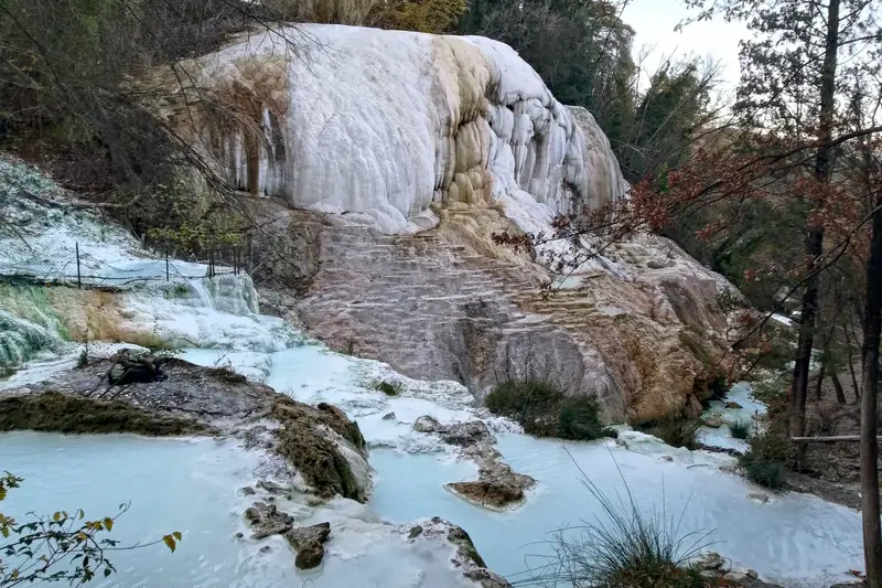 La cascade calcaire blanche de la Balena Bianca et les vasques d’eau bleu pâle du Fosso Bianco, au milieu d’un bois aux arbres dénudés, à Bagni San Filippo en Toscane.