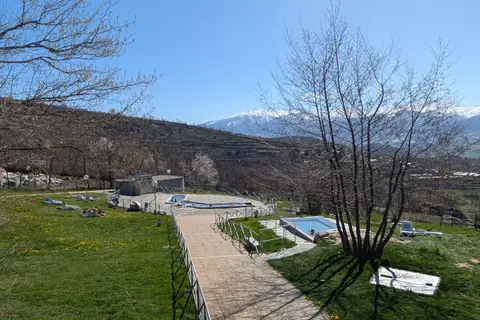 Les deux bassins extérieurs des Bains de Dorres vus d’en haut, au bout d’une allée dallée, entre des pelouses et des arbres sans feuilles, avec les sommets enneigés de la Cerdagne au fond.