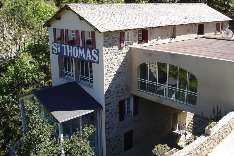 Le bâtiment des Bains de Saint-Thomas vu d’en haut : façade de pierre et d’enduit sous un toit de lauzes, grande enseigne « St THOMAS » bleue et blanche, verrières, au milieu des arbres.
