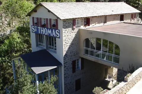 Le bâtiment des Bains de Saint-Thomas vu d’en haut : façade de pierre et d’enduit sous un toit de lauzes, grande enseigne « St THOMAS » bleue et blanche, verrières, au milieu des arbres.
