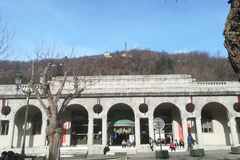 La façade des Bains du Couloubret en hiver : une galerie d’arcades en pierre claire, percée d’oculus, avec l’enseigne « Bains du Couloubret » au-dessus de l’entrée vitrée. Quelques personnes sont assises sur les marches, sous un versant boisé et un ciel bleu.
