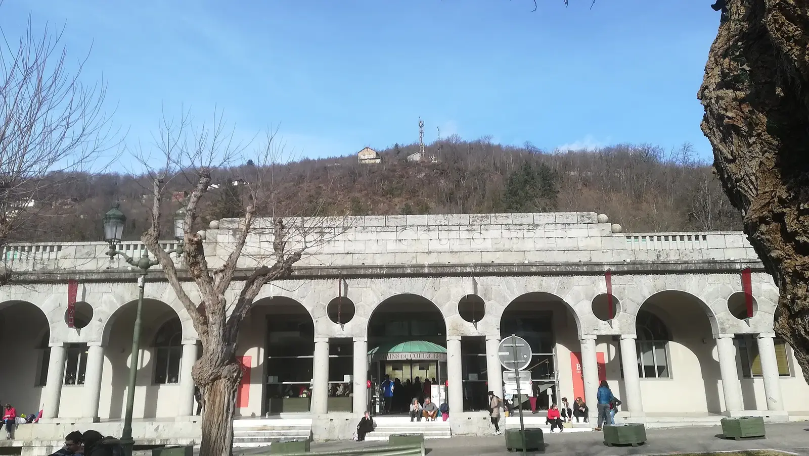 La façade des Bains du Couloubret en hiver : une galerie d’arcades en pierre claire, percée d’oculus, avec l’enseigne « Bains du Couloubret » au-dessus de l’entrée vitrée. Quelques personnes sont assises sur les marches, sous un versant boisé et un ciel bleu.