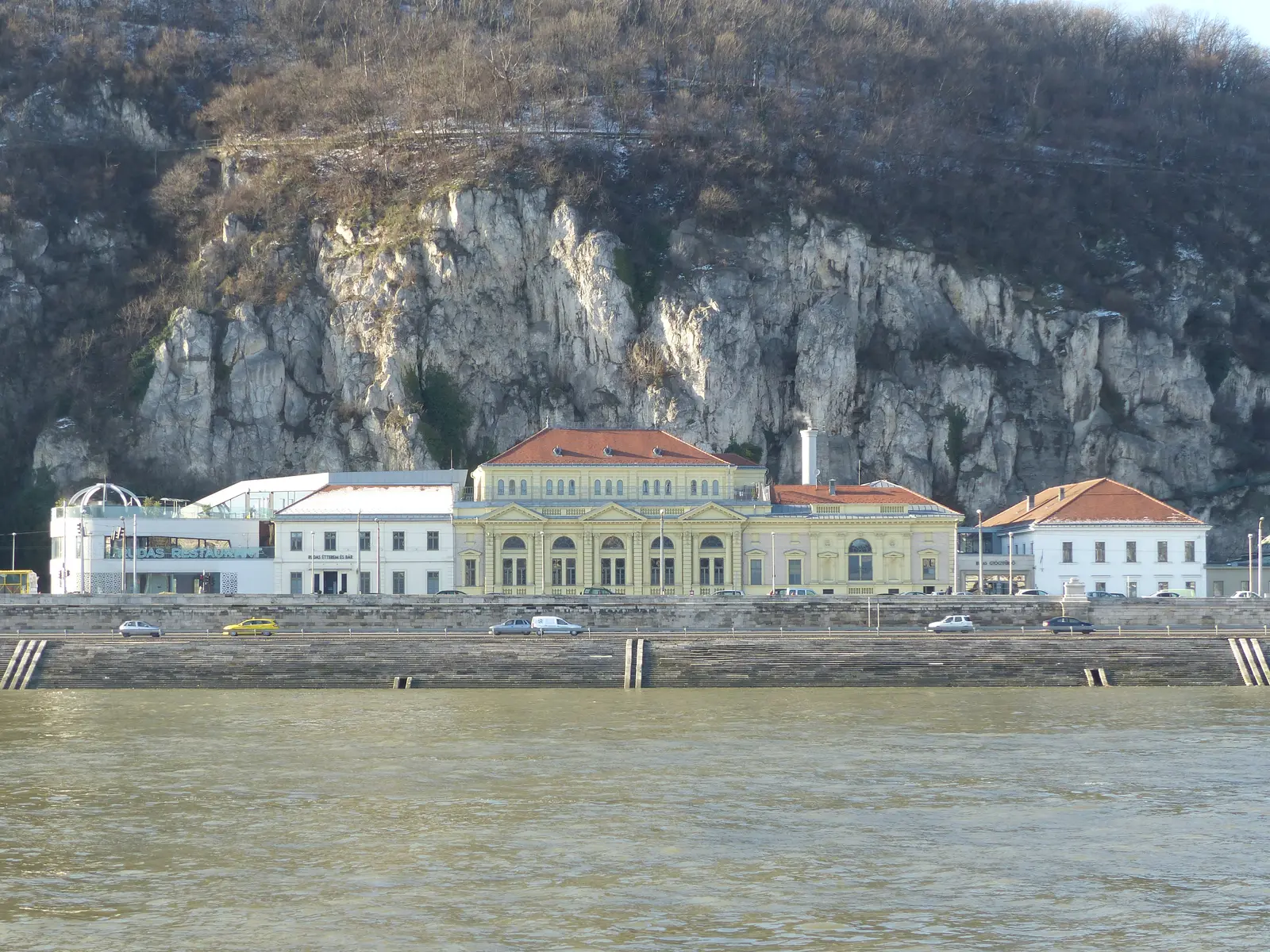 Façade néoclassique jaune et toits de tuiles rouges des bains Rudas, alignés le long du quai de Buda au pied de la falaise du mont Gellért, vus depuis l'autre rive du Danube par temps clair d'hiver.