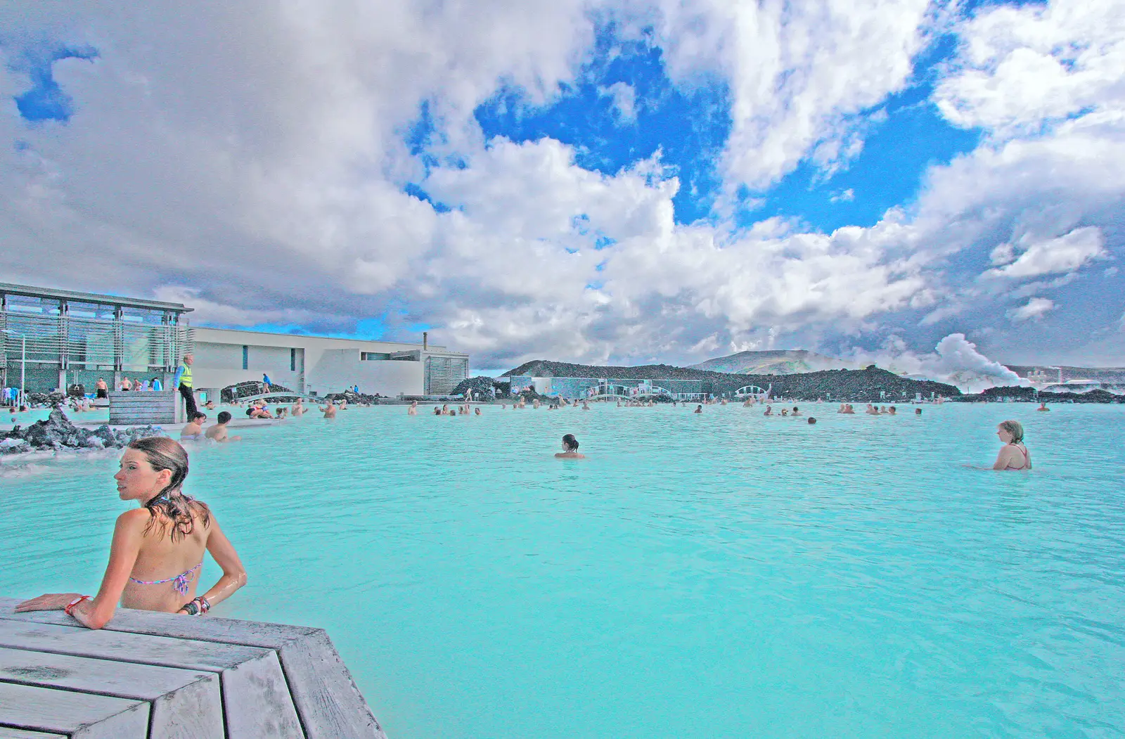 Une trentaine de baigneurs dans l’eau turquoise du lagon, devant le bâtiment d’accueil vitré et une crête de lave noire, sous un ciel nuageux.