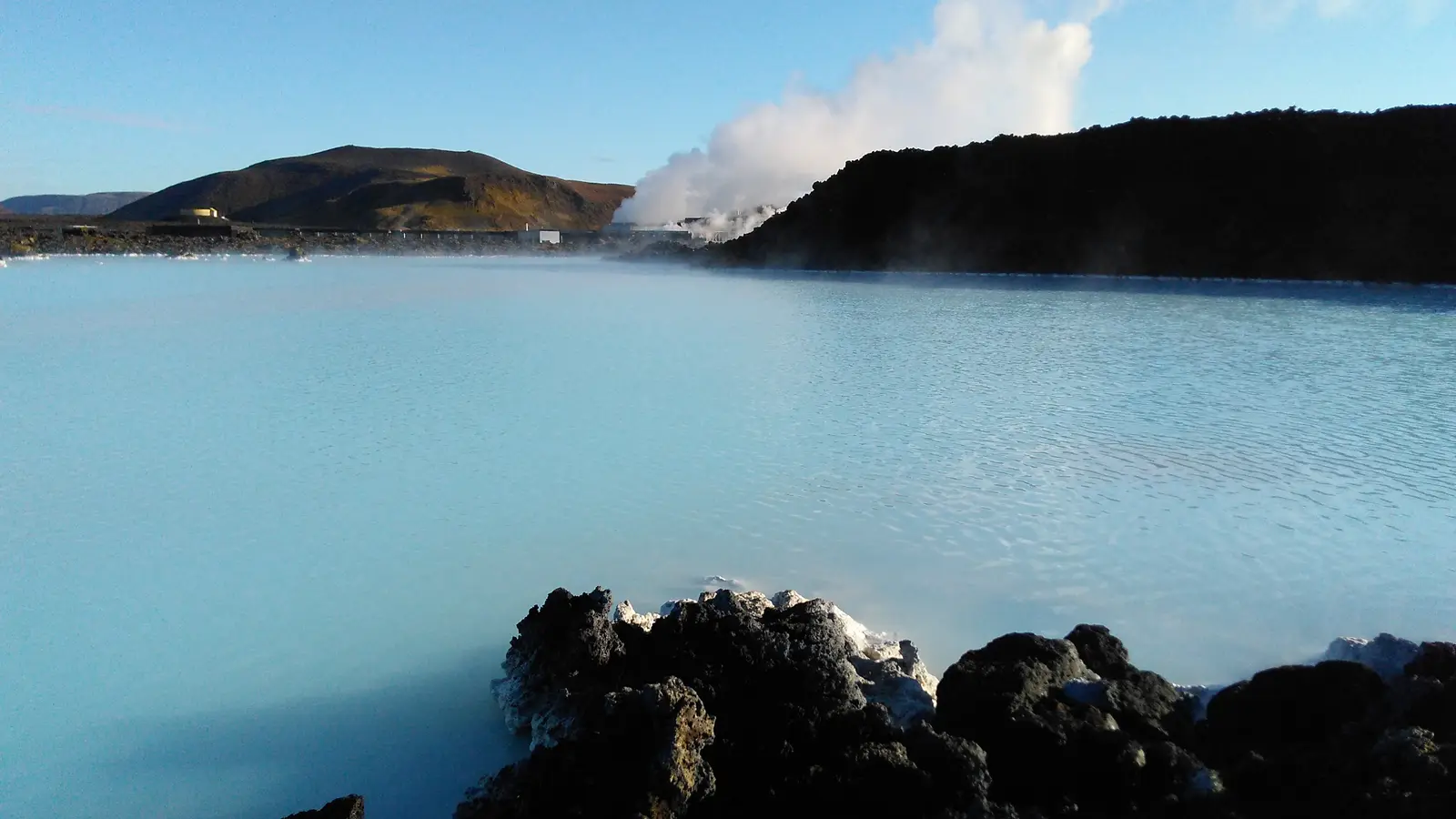 Le lagon du Blue Lagoon, eau bleu pâle et fumante entre des blocs de lave noire croûtés de dépôts blancs ; au fond, une colline brune et un panache de vapeur au-dessus d’une crête de lave.