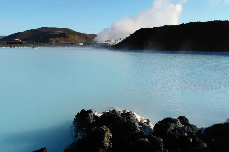 Le lagon du Blue Lagoon, eau bleu pâle et fumante entre des blocs de lave noire croûtés de dépôts blancs ; au fond, une colline brune et un panache de vapeur au-dessus d’une crête de lave.