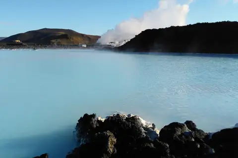 Le lagon du Blue Lagoon, eau bleu pâle et fumante entre des blocs de lave noire croûtés de dépôts blancs ; au fond, une colline brune et un panache de vapeur au-dessus d’une crête de lave.