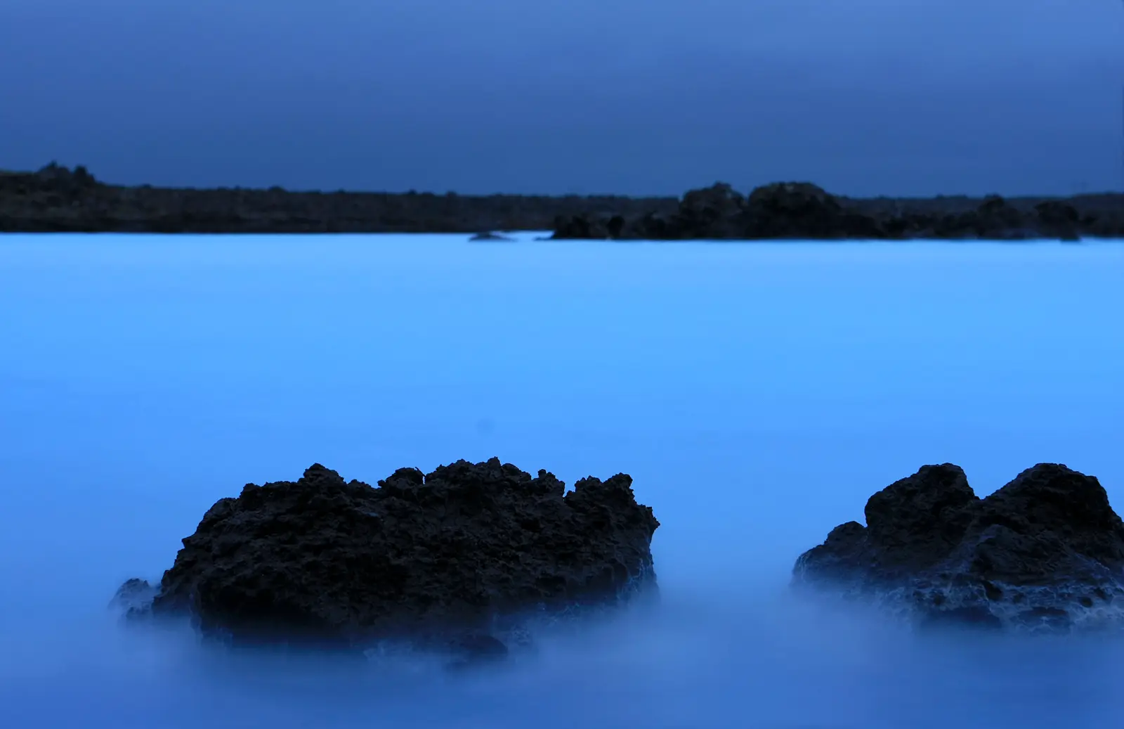 Deux blocs de lave sombres émergeant d’une eau bleu laiteux lissée par une pose longue, au crépuscule, devant une berge de lave basse.