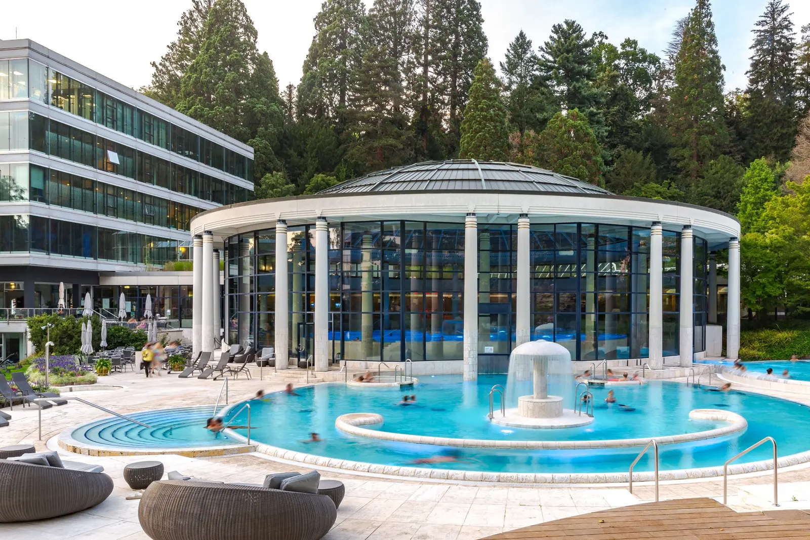 Le bassin extérieur de la Caracalla Therme au crépuscule : une eau turquoise où nagent quelques baigneurs, un jet central en forme de champignon, et derrière, la rotonde vitrée qui couvre le bassin intérieur, adossée à la forêt.