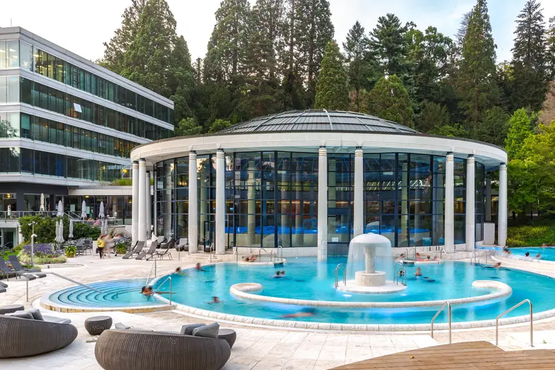 Le bassin extérieur de la Caracalla Therme au crépuscule : une eau turquoise où nagent quelques baigneurs, un jet central en forme de champignon, et derrière, la rotonde vitrée qui couvre le bassin intérieur, adossée à la forêt.