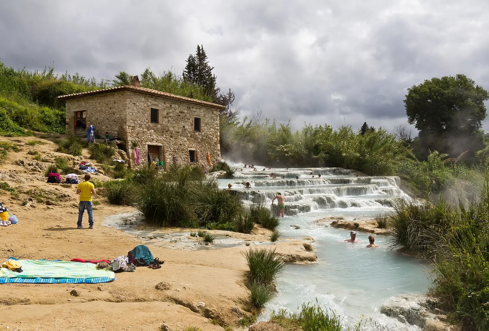 Le moulin de pierre des Cascate del Mulino au-dessus des gradins calcaires d'où monte la vapeur, avec quelques baigneurs au loin dans les vasques et des serviettes étendues sur la berge.