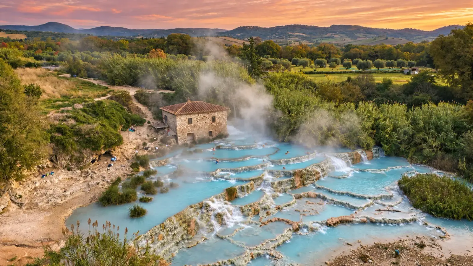 Vasques blanches étagées des Cascate del Mulino, remplies d'une eau bleu pâle d'où monte la vapeur, au pied du vieux moulin de pierre, avec les collines boisées de la Maremme sous un ciel de soleil couchant.