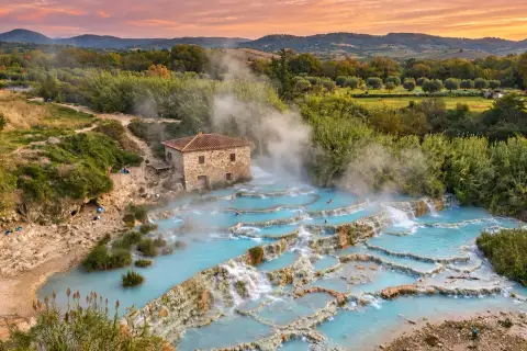 Vasques blanches étagées des Cascate del Mulino, remplies d'une eau bleu pâle d'où monte la vapeur, au pied du vieux moulin de pierre, avec les collines boisées de la Maremme sous un ciel de soleil couchant.