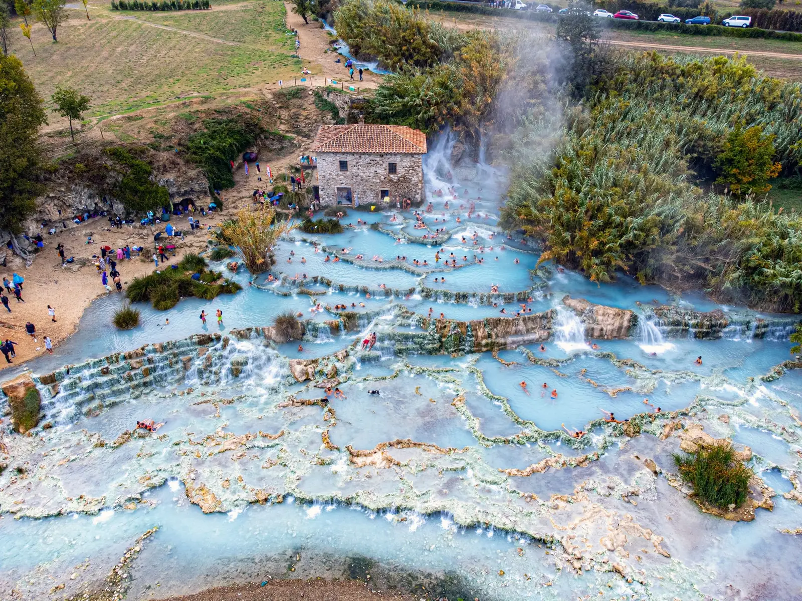 Vue aérienne des Cascate del Mulino en automne : des dizaines de baigneurs dans les vasques bleu pâle étagées devant le moulin de pierre, la vapeur au-dessus de la cascade et les prés de la Maremme autour.