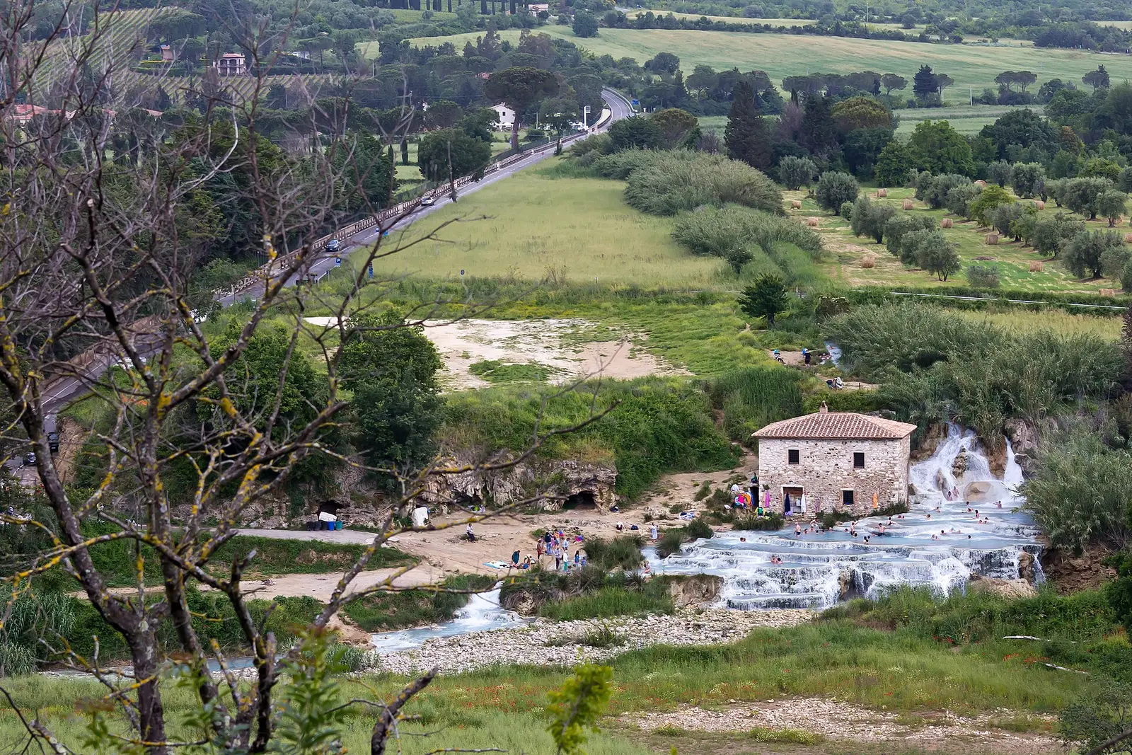 Le moulin de pierre et les gradins calcaires des Cascate del Mulino vus depuis la colline, au milieu des champs et des haies de la Maremme.
