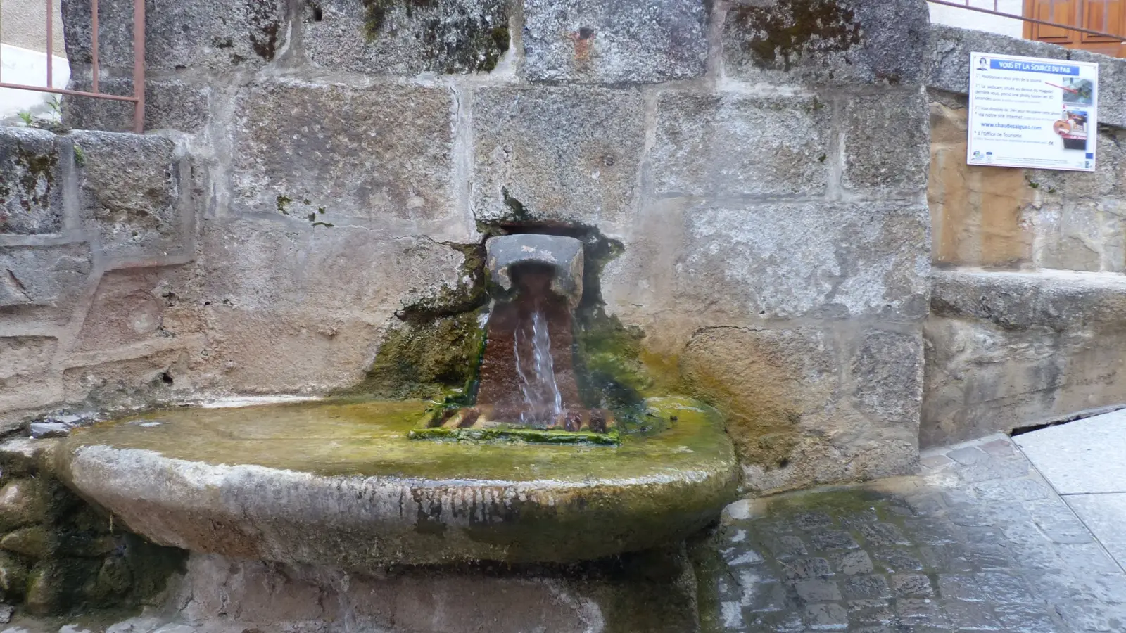 La source du Par à Chaudes-Aigues : l'eau thermale sort d'un bec de pierre scellé dans un mur de gros blocs de granit et tombe dans une large vasque de pierre, dont le fond est teinté d'ocre et de vert par les dépôts.