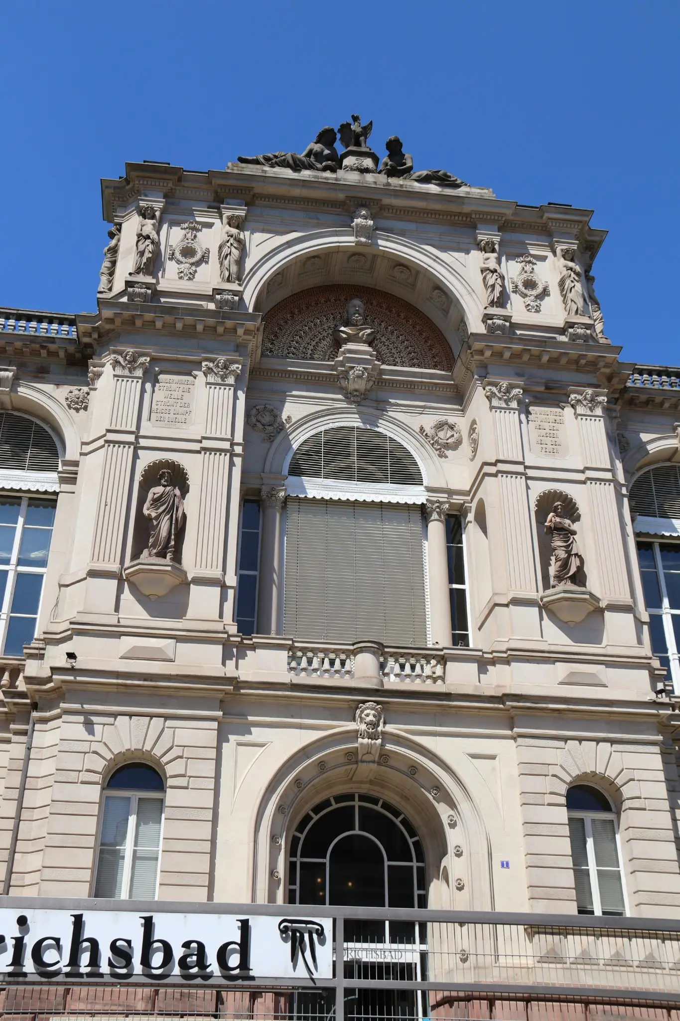 Façade de grès du Friedrichsbad à Baden-Baden, vue en contre-plongée : statues en niches, cariatides, plaques gravées et groupe sculpté au sommet, sous un ciel bleu.