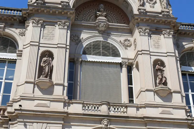 Façade de grès du Friedrichsbad à Baden-Baden, vue en contre-plongée : statues en niches, cariatides, plaques gravées et groupe sculpté au sommet, sous un ciel bleu.