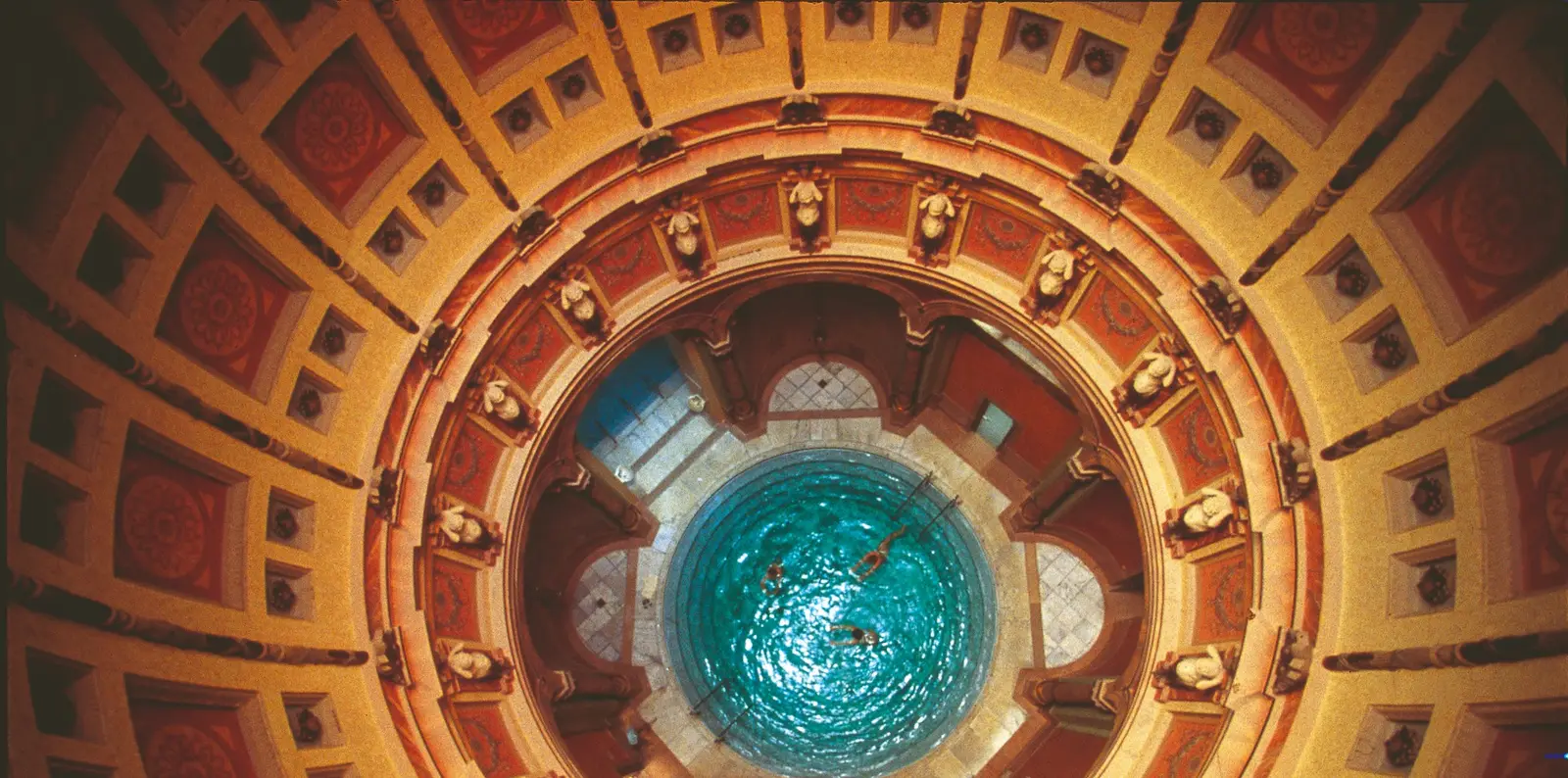 Vue plongeante sur la salle sous coupole du Friedrichsbad : le bassin circulaire d’eau turquoise au centre, entouré d’une couronne de statues et de caissons peints d’ocre et de rouge.