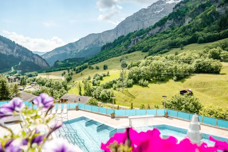Bassins extérieurs de Leukerbad Therme sur une terrasse fleurie, ouverts sur la vallée boisée et les parois rocheuses du Valais, en été.