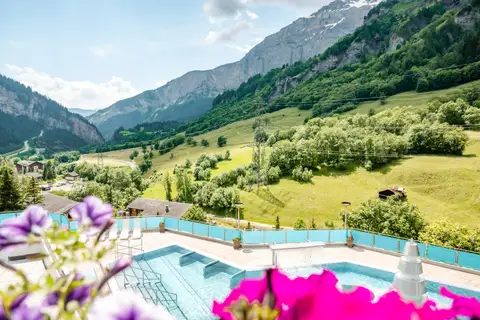 Bassins extérieurs de Leukerbad Therme sur une terrasse fleurie, ouverts sur la vallée boisée et les parois rocheuses du Valais, en été.