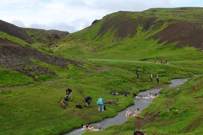 Baigneurs assis dans la rivière chaude de Reykjadalur, au fond d’une vallée verte encadrée de pentes herbeuses, avec des randonneurs sur la berge.