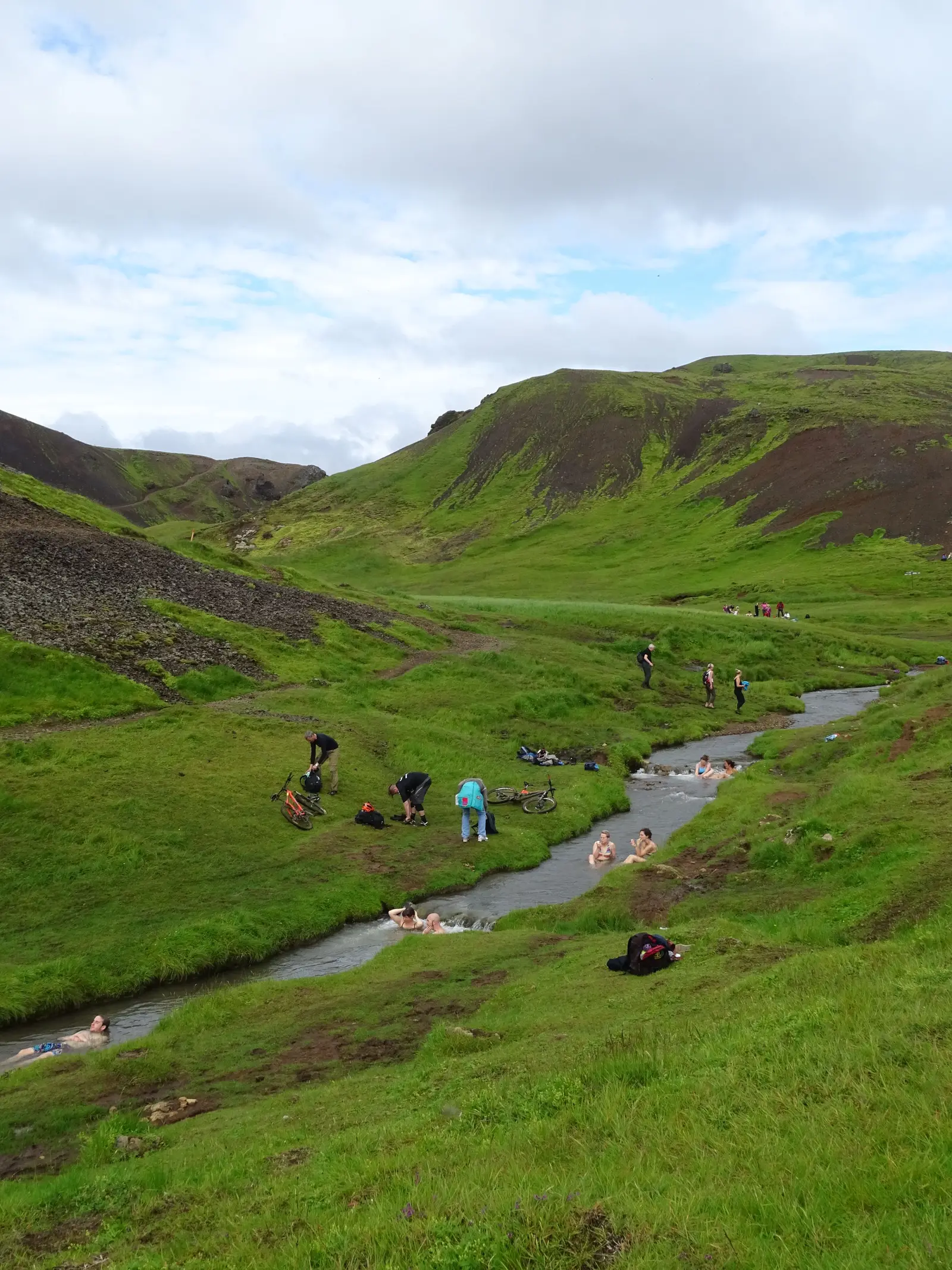 Baigneurs assis dans la rivière chaude de Reykjadalur, au fond d’une vallée verte encadrée de pentes herbeuses, avec des randonneurs sur la berge.