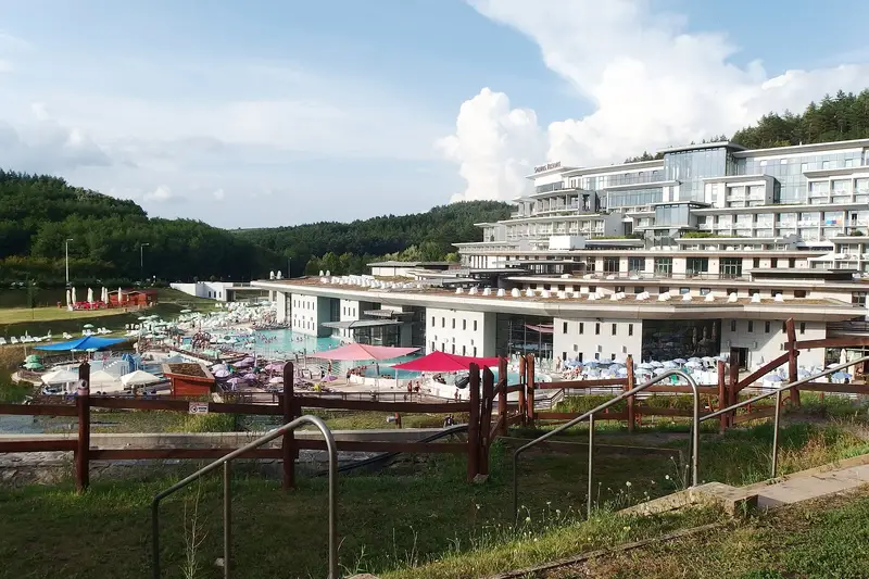 Vue plongeante sur des bassins en plein air bordés de parasols et de transats, devant un grand hôtel blanc en terrasses marqué « Saliris Resort », au pied de collines boisées.
