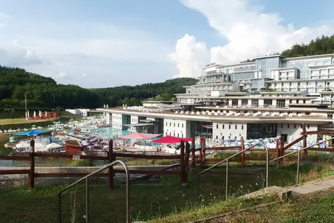 Vue plongeante sur des bassins en plein air bordés de parasols et de transats, devant un grand hôtel blanc en terrasses marqué « Saliris Resort », au pied de collines boisées.