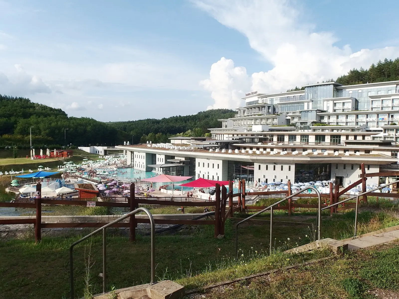 Vue plongeante sur des bassins en plein air bordés de parasols et de transats, devant un grand hôtel blanc en terrasses marqué « Saliris Resort », au pied de collines boisées.