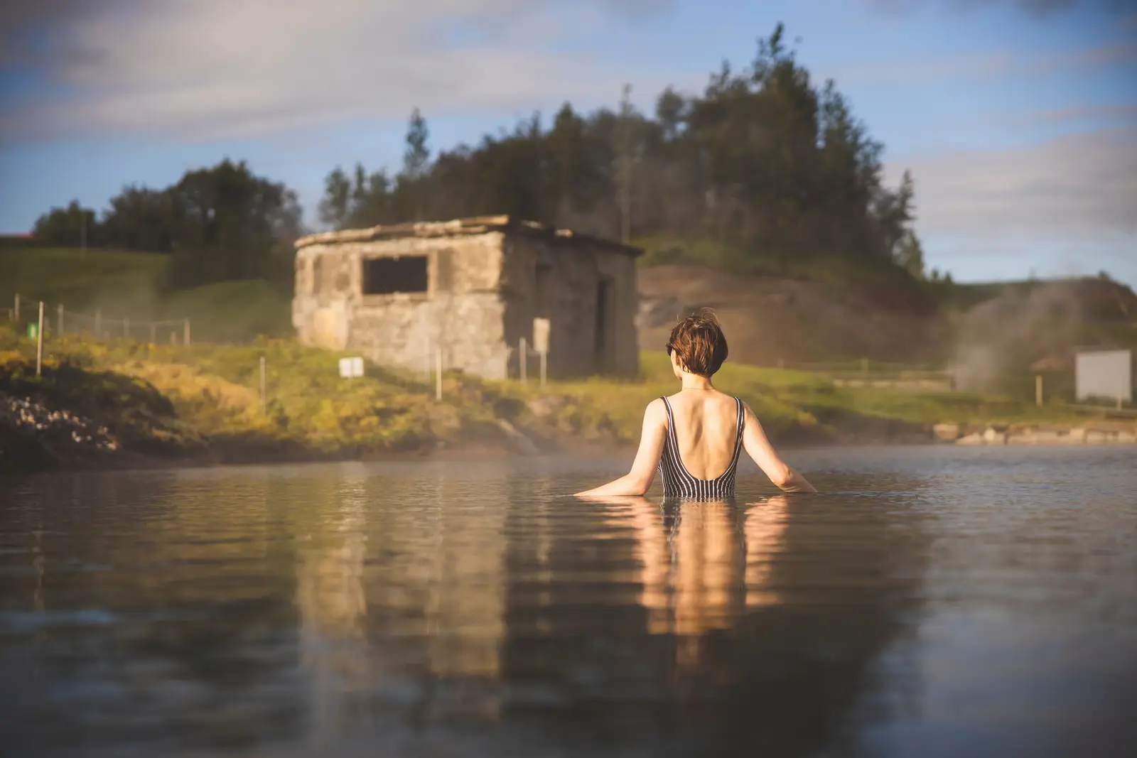 Une baigneuse en maillot rayé, vue de dos, dans l’eau du bassin jusqu’à la taille ; devant elle, une ancienne cabane de béton, de l’herbe et des arbres sous un ciel bleu, de la vapeur à droite.
