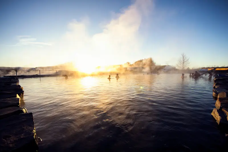 Le grand bassin au soleil bas d’hiver : une eau sombre et lisse bordée de dalles de pierre, quelques baigneurs au loin, de la vapeur qui monte dans la lumière, un arbre nu et des bâtiments à l’horizon.