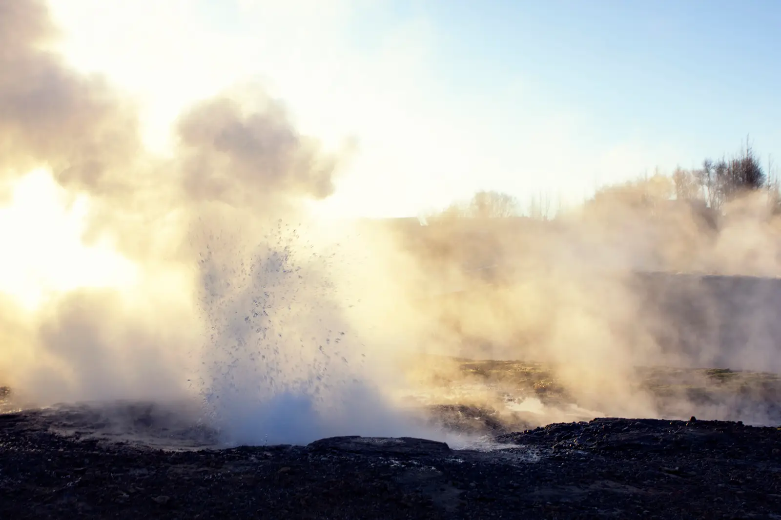 Une gerbe d’eau et de vapeur jaillit d’un sol sombre, à contre-jour, dans un nuage de vapeur doré par le soleil bas.
