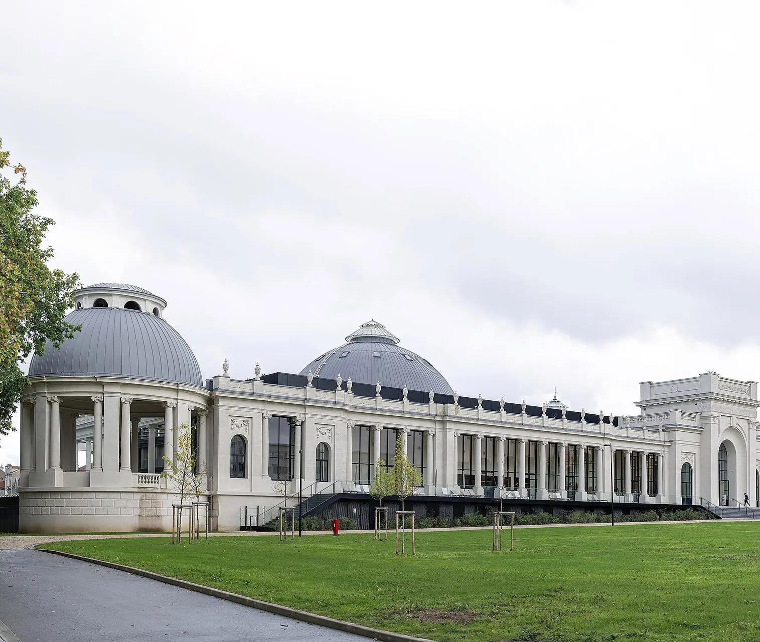 Les bâtiments historiques de Nancy Thermal, blancs et rénovés : un pavillon à colonnes coiffé d’un dôme gris, une longue galerie vitrée rythmée de colonnes, un second dôme et un pavillon d’entrée à arcade. Au premier plan, une pelouse plantée de jeunes arbres et une allée, sous un ciel couvert.
