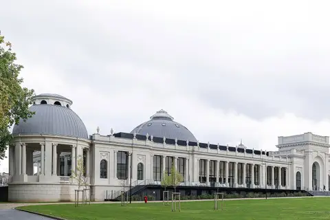 Les bâtiments historiques de Nancy Thermal, blancs et rénovés : un pavillon à colonnes coiffé d’un dôme gris, une longue galerie vitrée rythmée de colonnes, un second dôme et un pavillon d’entrée à arcade. Au premier plan, une pelouse plantée de jeunes arbres et une allée, sous un ciel couvert.