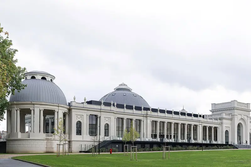 Les bâtiments historiques de Nancy Thermal, blancs et rénovés : un pavillon à colonnes coiffé d’un dôme gris, une longue galerie vitrée rythmée de colonnes, un second dôme et un pavillon d’entrée à arcade. Au premier plan, une pelouse plantée de jeunes arbres et une allée, sous un ciel couvert.