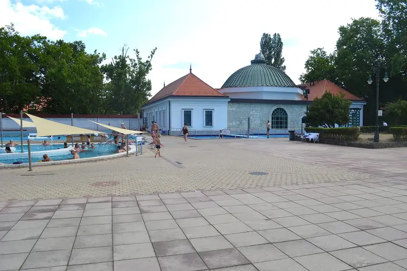 Un bassin en plein air sous des voiles d’ombrage, quelques baigneurs, une vaste esplanade dallée et, au fond, un pavillon blanc et en pierre coiffé d’une coupole verte, entre de grands arbres.