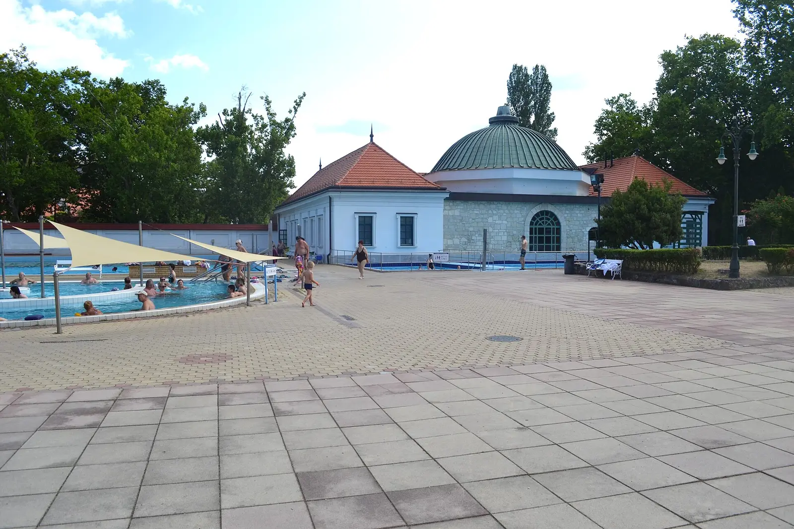 Un bassin en plein air sous des voiles d’ombrage, quelques baigneurs, une vaste esplanade dallée et, au fond, un pavillon blanc et en pierre coiffé d’une coupole verte, entre de grands arbres.
