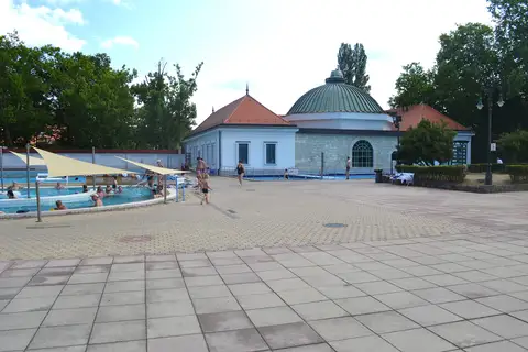 Un bassin en plein air sous des voiles d’ombrage, quelques baigneurs, une vaste esplanade dallée et, au fond, un pavillon blanc et en pierre coiffé d’une coupole verte, entre de grands arbres.