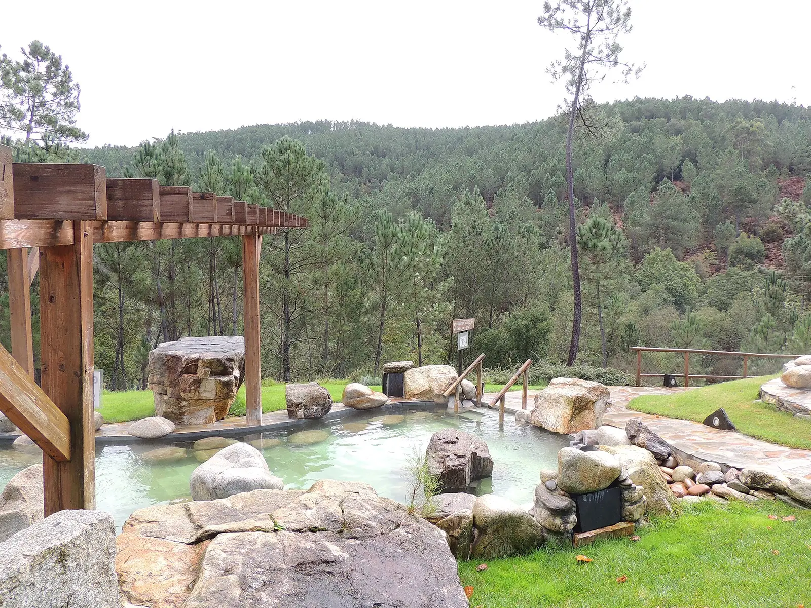 Un bassin d’eau verte semé de gros blocs de granite, vu à travers une pergola en bois, entre pelouse et dallage, devant une colline couverte de pins.