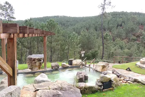 Un bassin d’eau verte semé de gros blocs de granite, vu à travers une pergola en bois, entre pelouse et dallage, devant une colline couverte de pins.