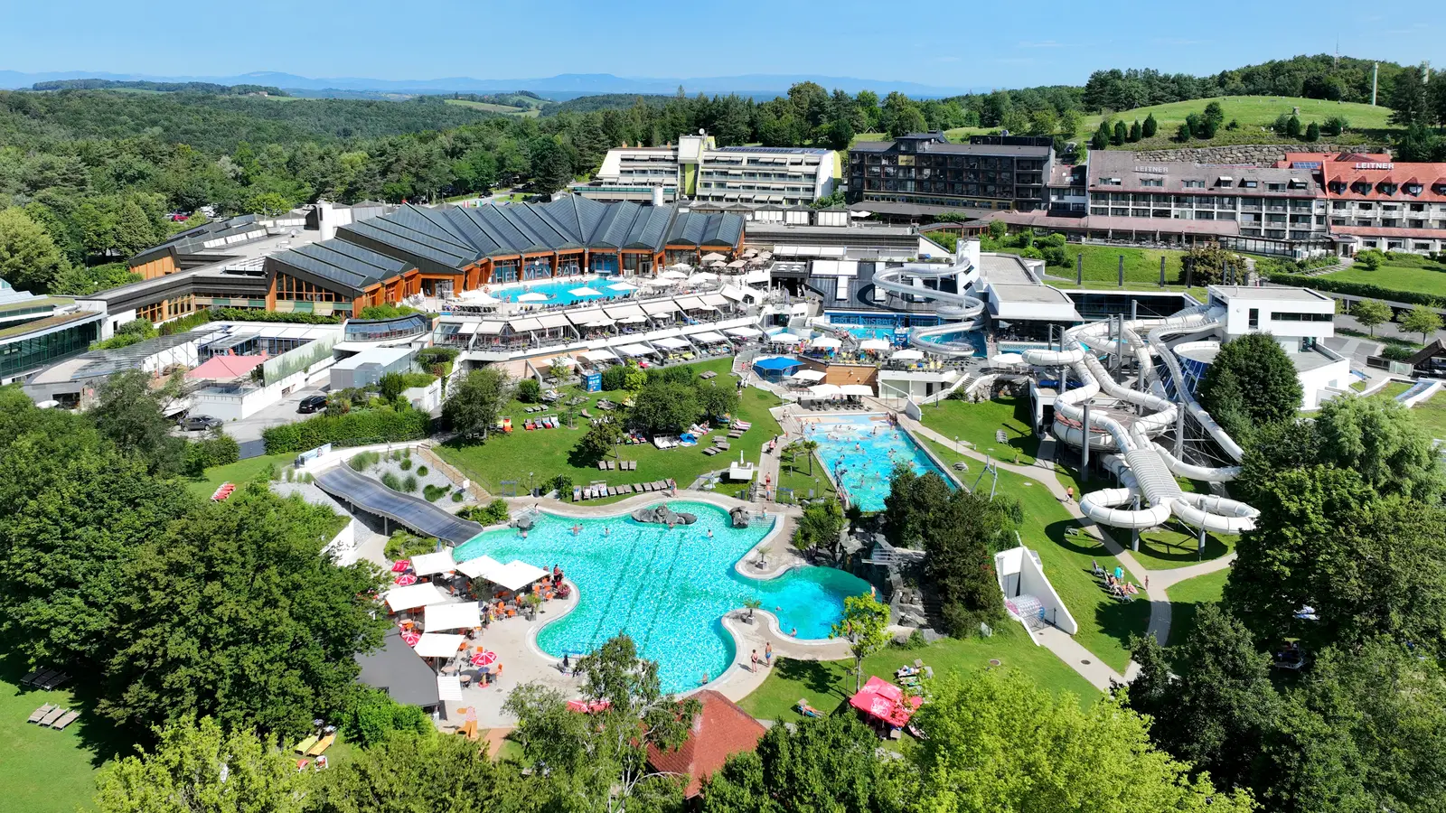 Vue aérienne d’un vaste complexe de baignade en été : un bassin extérieur turquoise aux contours arrondis entouré de parasols et de transats, un second bassin rectangulaire, des toboggans blancs enroulés, des halls aux toits de verre sombre et, au fond, des hôtels sur une colline boisée.