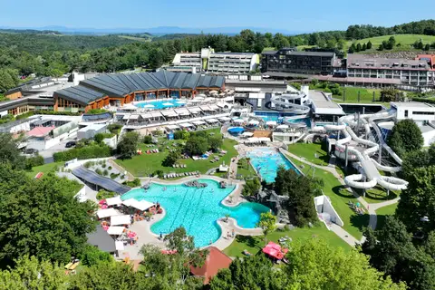 Vue aérienne d’un vaste complexe de baignade en été : un bassin extérieur turquoise aux contours arrondis entouré de parasols et de transats, un second bassin rectangulaire, des toboggans blancs enroulés, des halls aux toits de verre sombre et, au fond, des hôtels sur une colline boisée.