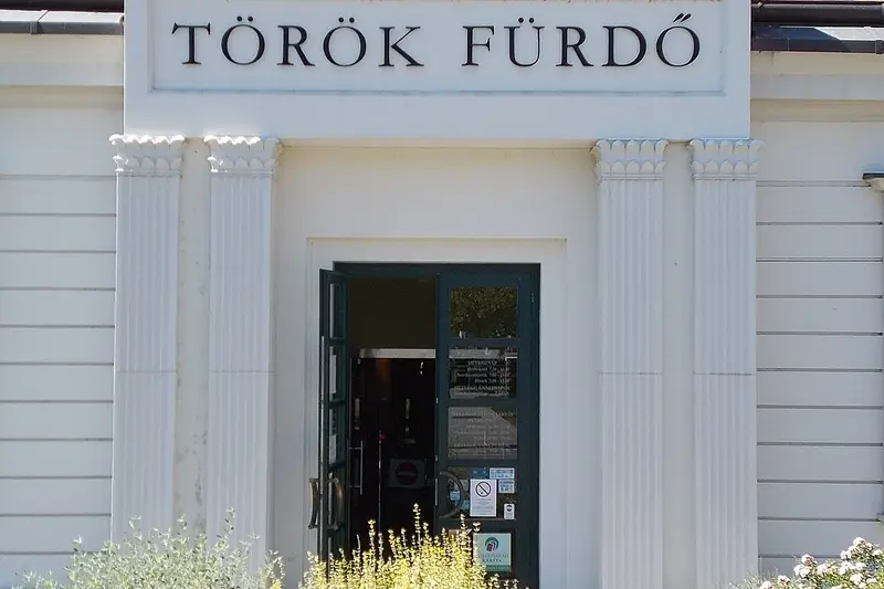 Façade blanche à pilastres sous un toit de tuiles orange, surmontée de l’inscription « TÖRÖK FÜRDŐ » ; la porte vitrée est ouverte, des massifs fleuris bordent le trottoir.